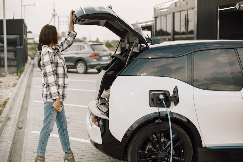 Woman on the electric cars charge station at daytime. Woman standing near opened trunk of her electrocar. Brunette woman wearing plaid jacket and jeans.